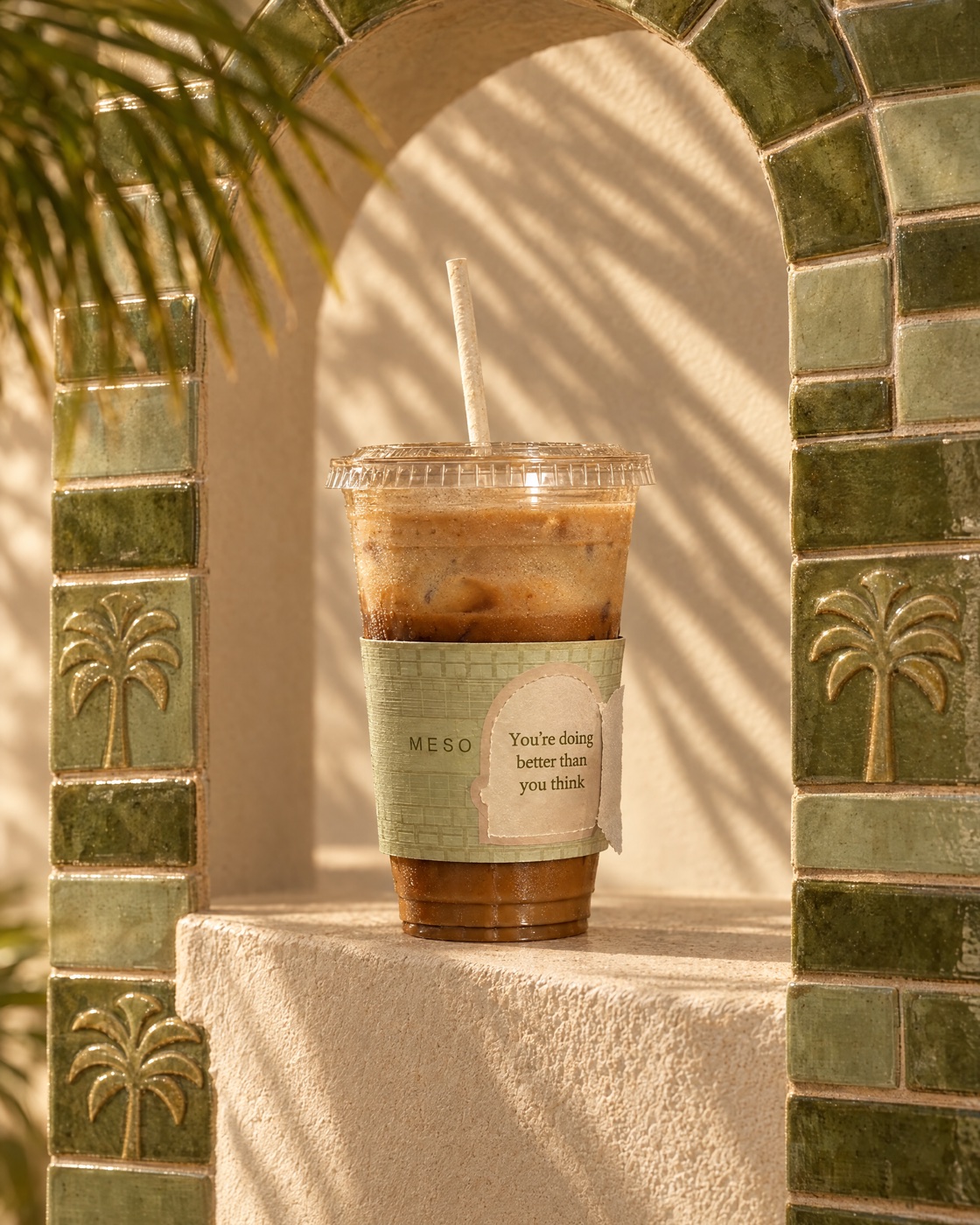 An iced latte in a Meso cup standing in an arch of olive glazed bricks with palm tiles, inspired by the Ishtar Gate